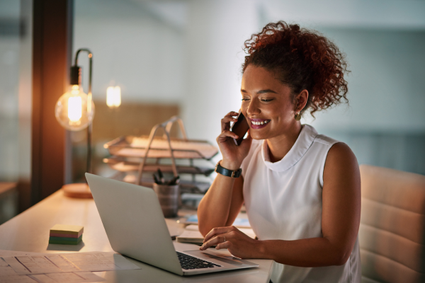 mujer sonriendo frente a un equipo portátil y sosteniendo un teléfono